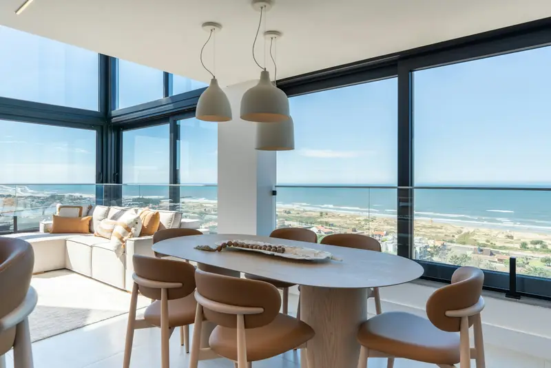 A dining table beside floor-to-ceiling corner glass, with the ocean beyond